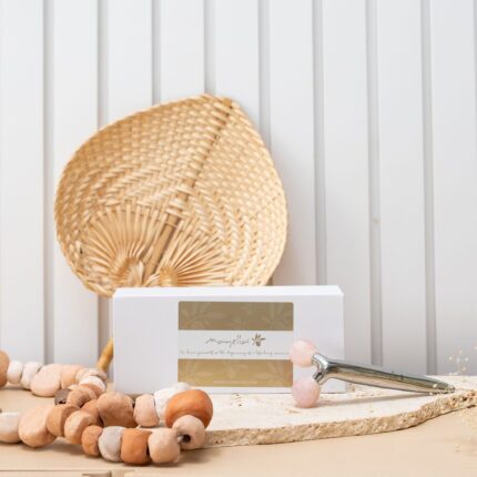 A cane woven fan in the background behind the white luxe box of Mayella Facial defining roller with two faceted rose quartz balls lying on a disc of cream natural stone. A string of natural toned wooden beads lies to the front left.